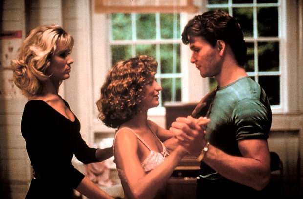 scene from dance training montage in dirty dancing where cynthia rhodes (penny) - wearing a black leotard - helps jennifer grey (baby) - wearing a pink bra and shorts - move the proper steps with patrick swayze (johnny) - wearing a gray shirt and black pants