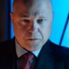 actor michael chicklis in image from fox series gotham, wearing a dark suit with a striped shirt and red tie and a grim expression on his face