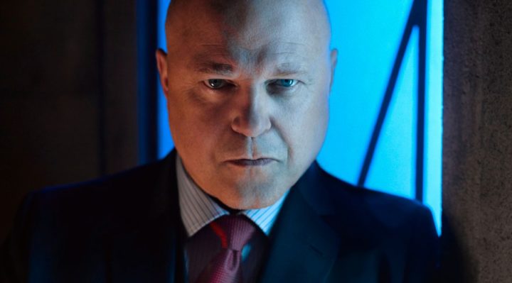 actor michael chicklis in image from fox series gotham, wearing a dark suit with a striped shirt and red tie and a grim expression on his face