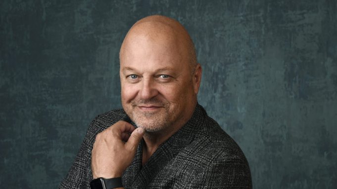 headshot of actor michael chicklis smiling at the camera and posing with his hand under his chin in front of a dark blueish gray background while wearing a gray houndstooth suit